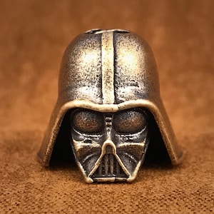 May include: A bronze-colored metal bead shaped like Darth Vader's head. The bead has a detailed design of Darth Vader's helmet, including the iconic black visor and breathing apparatus.