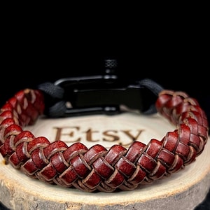 Op de afbeelding: Gevlochten bordeaux leren armband met een zwarte sluiting. De armband wordt getoond op een houten oppervlak met het Etsy-logo. Het leer is geweven in een dik, getextureerd patroon, en de sluiting heeft een matzwarte afwerking.