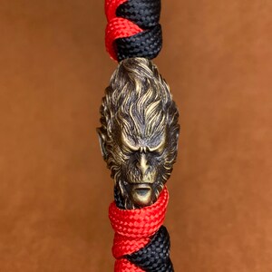 Pode incluir: Um bracelete de paracord preto e vermelho com um amuleto de macaco de latão detalhado. O amuleto de macaco tem uma aparência texturizada, quase peluda.