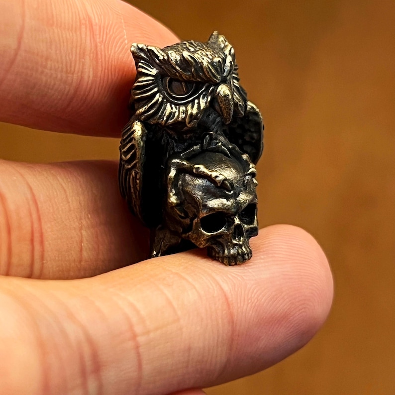 Puede incluir: Una figura de b&uacute;ho de color bronce posada sobre una calavera. El b&uacute;ho tiene plumas detalladas y ojos grandes y redondos. La calavera tiene dientes y cuencas de los ojos visibles. La figura se sostiene entre los dedos, lo que sugiere su peque&ntilde;o tama&ntilde;o.