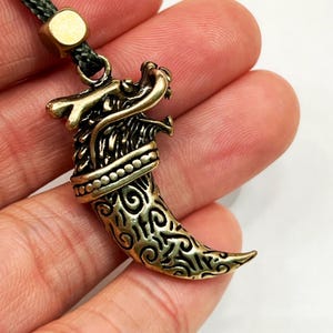 Könnte beinhalten: Bronzefarbener Anhänger in Form einer Drachenkralle. Der Anhänger ist detailliert mit einem Drachenkopf und kunstvollen Mustern auf der Kralle gestaltet. Er hängt an einer dunkelgrünen Kordel mit einem goldfarbenen Verschluss.