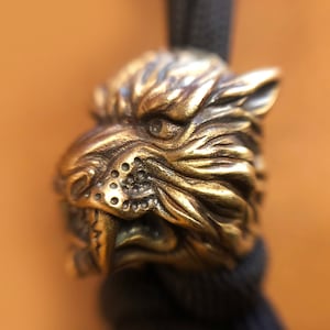 Puede incluir: Un amuleto de metal dorado en forma de cabeza de lobo gruñendo. El amuleto está detallado con pelaje texturizado y dientes afilados.