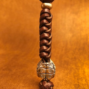 May include: A brown leather cord with a braided design and a gold metal turtle charm. The turtle charm has Chinese characters on its shell.