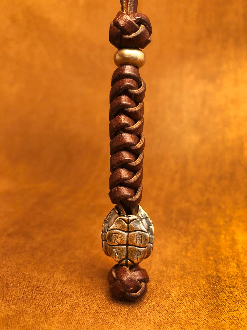 May include: A brown leather cord with a braided design and a gold metal turtle charm. The turtle charm has Chinese characters on its shell.