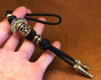 May include: A black lanyard with a bronze-colored Spartan helmet charm and decorative beads. The lanyard is made of a woven black cord and features two decorative end pieces. The charm is a detailed depiction of a Spartan helmet.