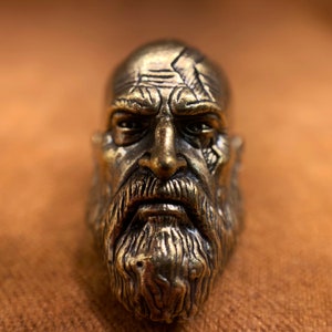 May include: A detailed bronze sculpture of a man's face with a beard. The sculpture has a weathered look and features a furrowed brow and a stern expression.