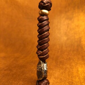 May include: A brown leather cord with a braided knot at the top and a brass bead. The cord is twisted and has a brass turtle charm at the bottom.
