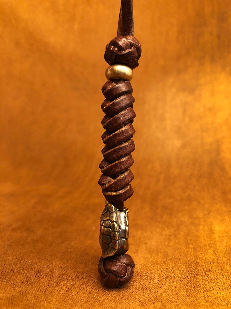 May include: A brown leather cord with a braided knot at the top and a brass bead. The cord is twisted and has a brass turtle charm at the bottom.