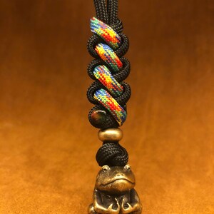 May include: A black and multi-colored lanyard with a small brass frog charm. The lanyard has a loop at the top for attaching to a key ring or bag.