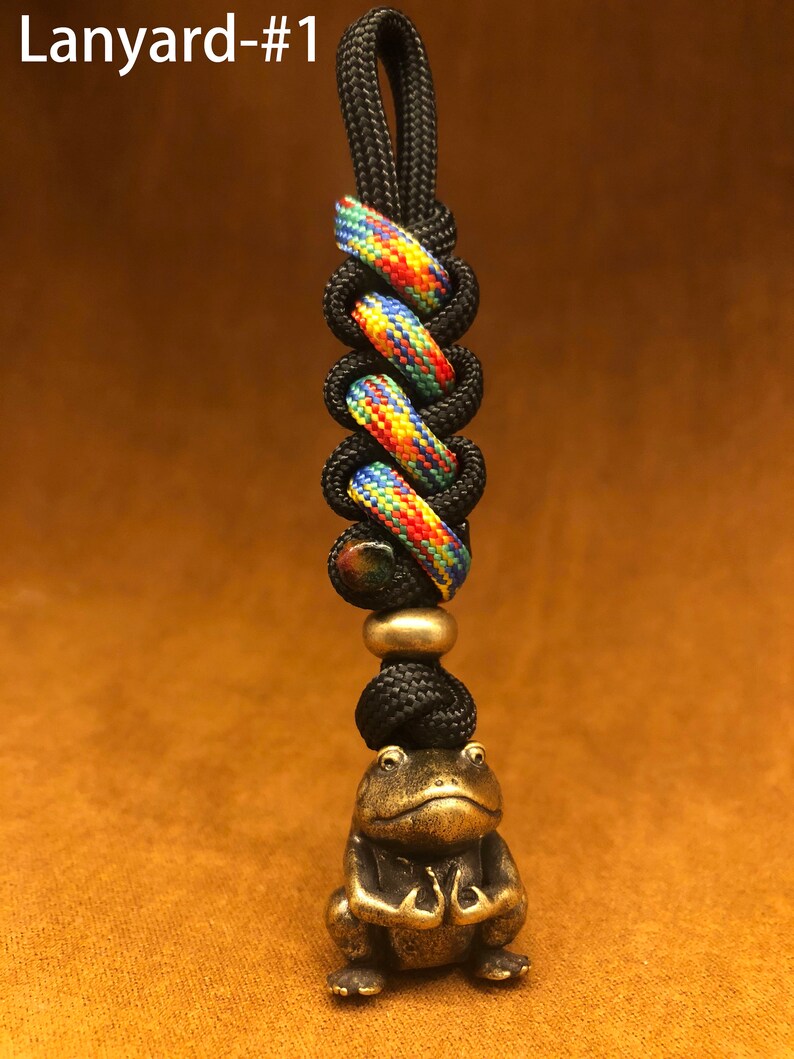 May include: A black and multi-colored lanyard with a small brass frog charm. The lanyard has a loop at the top for attaching to a key ring or bag.