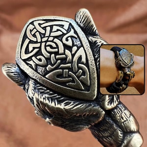 May include: A bronze-toned metal bracelet featuring a detailed wolf head clasp with a shield-shaped plate engraved with an intricate Celtic knot pattern. The bracelet is constructed from a black woven cord, showcasing the metallic wolf head design.
