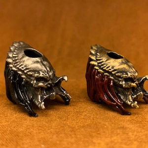 Pode incluir: Duas contas em forma de crânio de Predator, uma com acabamento preto e prata e a outra com acabamento dourado e vermelho.