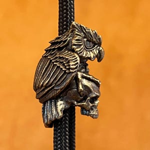 May include: A bronze-colored bolo tie pendant featuring an owl perched atop a skull. The owl has detailed feathering and a focused gaze. The pendant is attached to a black braided cord, set against an orange background.