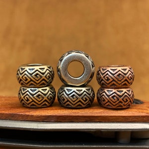 Op de afbeelding: Drie gestapelde metalen kralen met een geometrisch patroon. De bovenste kraal is zilver, de middelste kraal is goud en de onderste kraal is koper.