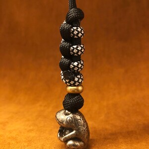 May include: A silver frog charm with a black and white paracord lanyard. The lanyard has a brass bead.