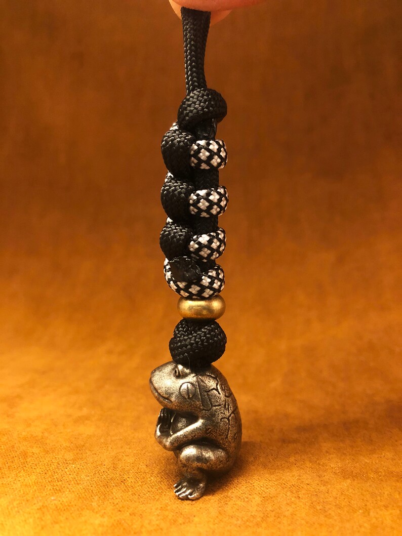 May include: A silver frog charm with a black and white paracord lanyard. The lanyard has a brass bead.