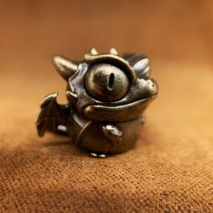 May include: A small, bronze-coloured dragon figurine with a single large eye and a toothy grin. The dragon has a small wing on its back and is standing on a brown surface.