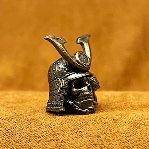 Könnte beinhalten: Ein detaillierter, bronzefarbener Schädel mit einem Samurai-Helm. Der Helm weist aufwendige geprägte Muster und ein dekoratives Wappen auf. Die Augenhöhlen und die Nasenhöhle des Schädels sind sichtbar.