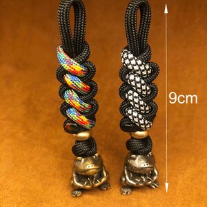 May include: Two black paracord keychains with different patterns, each with a small brass frog charm. The keychains are 9 cm long.