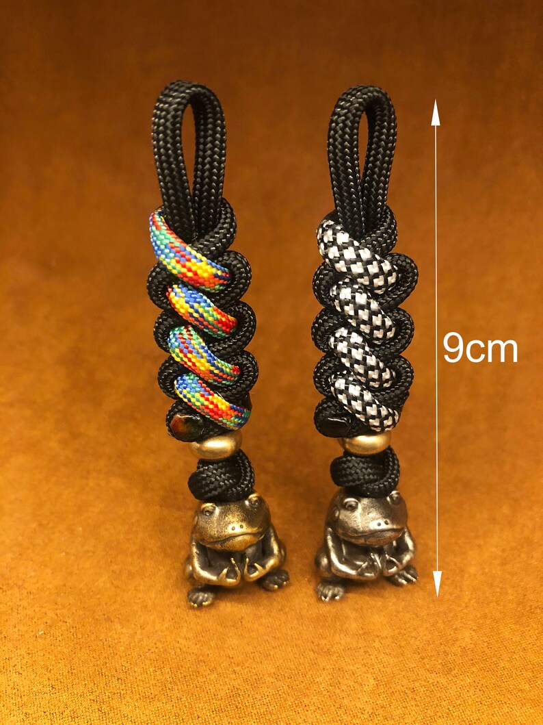 May include: Two black paracord keychains with different patterns, each with a small brass frog charm. The keychains are 9 cm long.