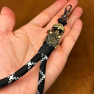 May include: A black and white braided cord lanyard featuring a metallic bronze-toned charm depicting a bearded figure with intricate knotwork patterns. The lanyard includes a black metal swivel lobster clasp at the top for secure attachment.