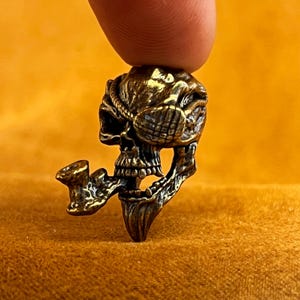 May include: A detailed bronze-colored skull pendant with an eye patch and a pipe. The skull has intricate details, including a rope around the head and a detailed jawline. The pendant is held by a finger against a yellow background.