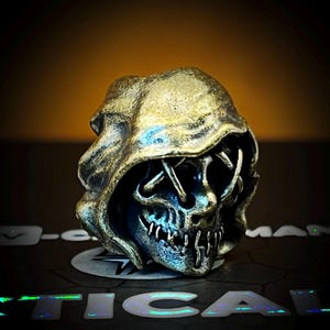 May include: A metallic skull ring with a hooded design. The ring features a detailed skull with stitched-up eyes and a menacing grin. The ring has a gold and silver finish, with a dark background.