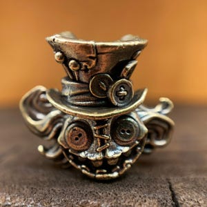 Peut inclure: Une figurine en laiton d'un personnage fantaisiste avec un chapeau haut de forme, des coutures cousues et de grands yeux en boutons. Le personnage a un large sourire et semble être fait de métal.