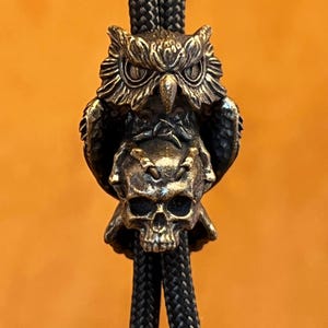 Puede incluir: Un colgante de corbata bolo en tono bronce que presenta un b&uacute;ho posado sobre una calavera. El b&uacute;ho tiene plumas detalladas y ojos penetrantes. La calavera tiene dientes y cuencas de los ojos visibles. El colgante est&aacute; suspendido de un cord&oacute;n trenzado negro, creando una est&eacute;tica g&oacute;tica o motera.
