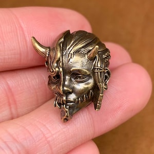 Peut inclure: Une sculpture en laiton d'un visage avec des cornes, divisé en deux pour révéler un crâne en dessous. La sculpture est détaillée et complexe, avec une surface texturée.