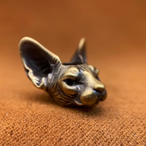 以下が含まれることがあります： スフィンクス猫の頭部の小さなブロンズ彫刻。彫刻は非常に詳細で、毛皮と顔の特徴がはっきりとわかります。