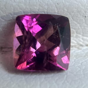 May include: A faceted, square-cut gemstone with a vibrant pink hue. The gem's surface reflects light, creating a play of color and depth. The gemstone is set against a neutral background.