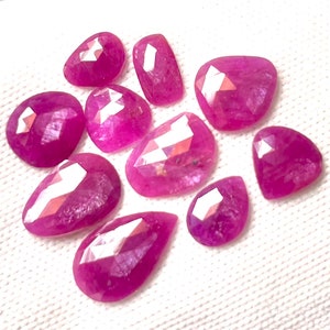 Puede incluir: Una colección de piedras preciosas de rubí rosa intenso facetadas en varias formas, incluyendo ovaladas, de pera e irregulares. Las gemas se exhiben sobre una superficie blanca, mostrando sus superficies pulidas y claridad interna.