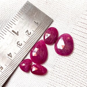 Può includere: Cinque pietre preziose rosa a forma di pera, ciascuna misura circa 1 cm di lunghezza, sono disposte su una superficie di tessuto bianco. Un righello di metallo è visibile sullo sfondo.