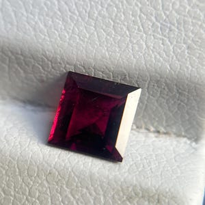 May include: A rectangular-cut, deep red gemstone with multiple facets. The gem has a polished surface and is set against a textured, off-white background. The gem's colour is a rich, dark red, with hints of purple.