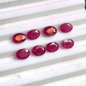 May include: Nine oval-shaped, red gemstones arranged in two rows on a white surface.