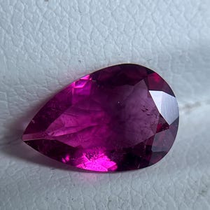 May include: A pear-shaped gemstone in a deep magenta colour. The cut of the gem reveals multiple facets that reflect light, creating a sparkling effect. The stone is set against a neutral background, highlighting its colour and shape.