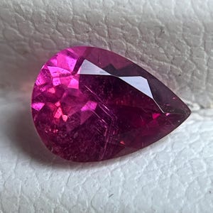 May include: A faceted, pear-shaped gemstone in a vibrant magenta hue. The gem's surface reflects light, showcasing its internal structure and cut. The stone is set against a textured, off-white background, highlighting its colour and brilliance. This gemstone is suitable for jewellery making.