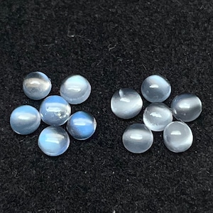 Op de afbeelding: Een verzameling ronde, witte maansteen cabochons met een lichte blauwe iriserende glans. De stenen zijn in twee groepen gerangschikt op een zwart oppervlak.