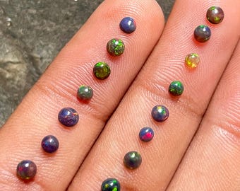 May include: Assortment of round, black opal gemstones with green, blue, and red iridescent flashes. The opals are displayed on a person's fingers.