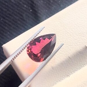 Puede incluir: Una piedra preciosa en forma de pera con un tono rojo intenso y un ligero tinte púrpura. La piedra preciosa se sostiene con unas pinzas sobre un fondo blanco.
