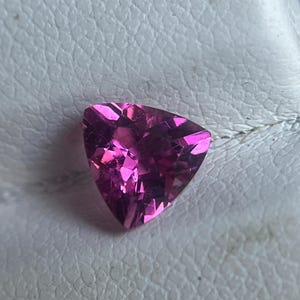 May include: A faceted, triangular-shaped gemstone with a vibrant pink hue. The gem's multiple facets reflect light, creating a sparkling effect. The gemstone is set against a textured, off-white surface.