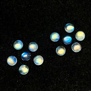 May include: Ten round, iridescent moonstone cabochons in various shades of blue and white. The stones are arranged in a scattered pattern on a black background.