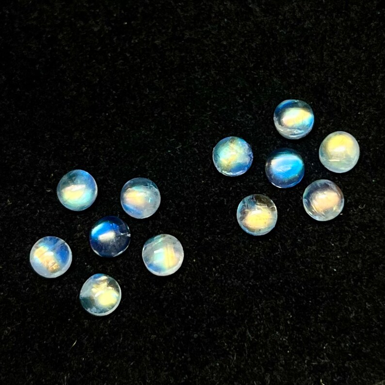 May include: Ten round, iridescent moonstone cabochons in various shades of blue and white. The stones are arranged in a scattered pattern on a black background.