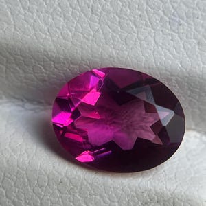 May include: An oval-cut, faceted gemstone in shades of deep magenta and fuchsia. The gem displays a star-like pattern within its depths, with sharp, angled facets catching the light. The stone is set against a textured, off-white surface.