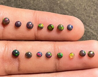 May include: A collection of 14 round, black opal gemstones with green and red flashes. The gemstones are arranged in two rows on a finger.