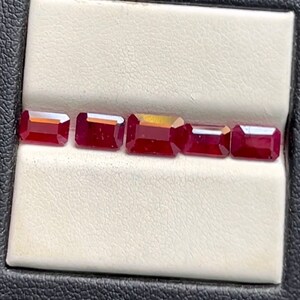 May include: Five red rectangular gemstones are displayed in a row on a white surface. The gemstones are all the same size and shape, and they are arranged in a straight line.