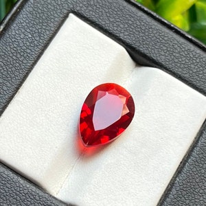 May include: A pear-shaped red gemstone with facets, set in a white display box.
