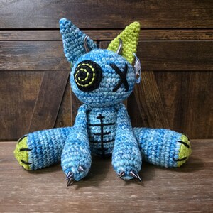 May include: A handmade blue crochet doll with black and yellow accents. The doll has a spiral eye, an X for an eye, and metal spikes on its feet. The ears are green and the doll has metal horns and a fish earring. The doll is sitting on a wooden surface.