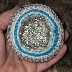 May include: A handmade, round, crocheted item with a geode-like design. The outer layers are gray and white yarn, with a turquoise blue band. The center is filled with small, shiny, silver beads.